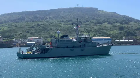 A warship on the water sailing to the right with a union jack visible and a Ukraine Navy flag also visible with some green hills in the background
