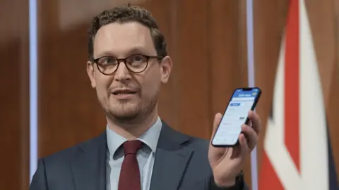 PA Media Darren Jones, in dark rimmed glasses and a blue suit with red tie, holds up a smartphone at a Downing Street press conference