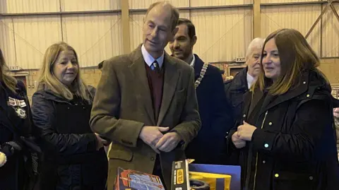 The Duke of Edinburgh stands amongst five people in front of trolleys of children's' toys. He is wearing a green blazer, burgundy sweater and white shirt. Those around him are wearing dark coloured coats.