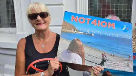 Alison Corlett has fair hair tied back and a fringe. She is wearing a black sleeveless top and is holding a NOT4IOM placard depicting Laxey breach with dozens of turbines on the coast line.