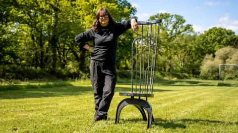 Urdd Woman in field leaning on a chair. She is wearing black trousers and a black long sleeve jumper. She has black glasses on.