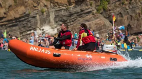 Stephen Duncombe/RNLI A picture of a Bude lifeboat. It is orange in colour and has two people wearing red life jackets.