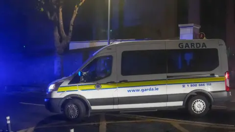 A white Garda van with a yellow stripe along its side pulls out onto a main road on a dark night.
