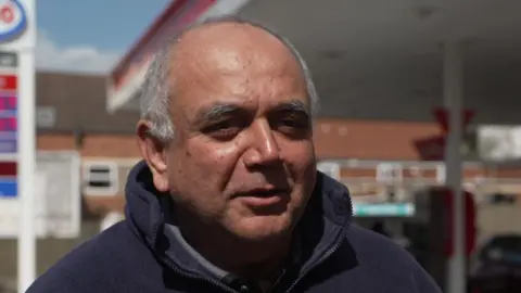 Shailesh Parekh - a clean shaven bald man with short grey hair at the sides of his head is standing in front of a petrol station.