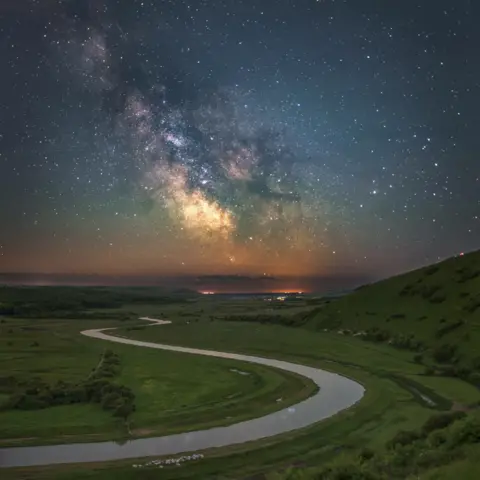 Jamie Fielding/South Downs National Park The Milky Way can be seen in a colourful night sky. The image shows a winding road snaking through a landscape of green. 