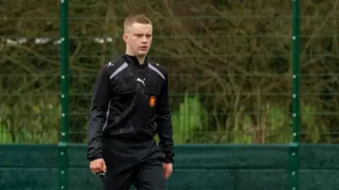 Jen Howell RJ refereeing a football game. He is walking along the pitch while wearing a black sports jacket and black trousers.