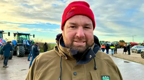 Linsey Smith/BBC Head and shoulders shot of Ed Pritchard. He is wearing a brown jacket and a red beanie hat. There are farm vehicles and people in the background.