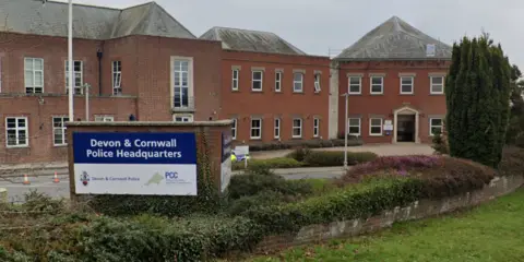 The Devon and Cornwall Police Headquarters building is a series of adjoining red brick, two-storey buildings. There are many windows and the buildings have grey slate roofs. There is a sign at the front which reads "Devon and Cornwall Police Headquarters" and bears the force's logo which is a map of Devon and Cornwall. There is a planted flowerbed in the foreground and a lawn.