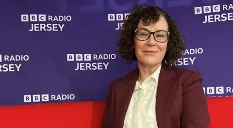BBC Carmel Corrigan wearing a burgundy jacket over a cream blouse. She has dark curly hair and broad glasses