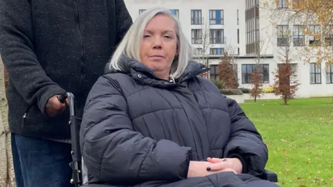 Karen James wearing a puffa jacket and sitting in her wheelchair on a grassy area in front of a block of flats