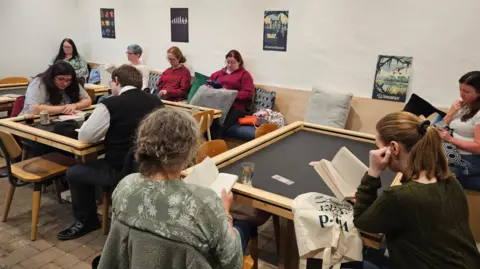 A group of nine people read books, on chairs and benches scattered with cushions.