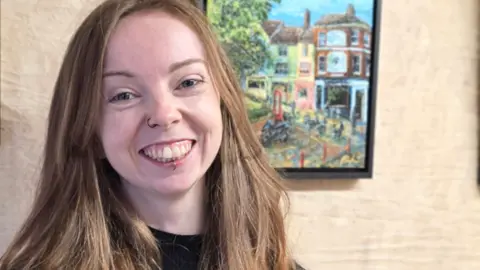 Lauren Pincher is standing inside a shipping container which has three framed pieces of her artwork behind her. She has long auburn hair and is wearing a black roll neck long sleeved T-shirt. She is looking directly at the camera and smiling.