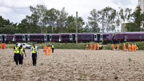EPA/Shutterstock Two purple, crashed trains in the background, with police and railway workers in orange standing in the field in front of the tracks.