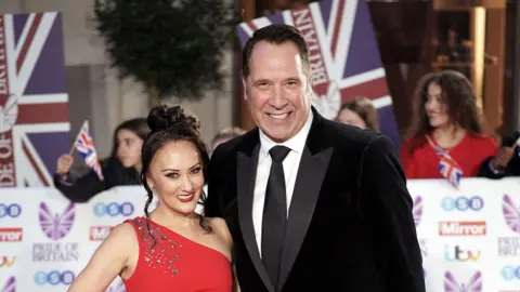AP Frankie and her husband David smiling on the red carpet at the Pride of Britain awards. Frankie is in a red dress over one shoulder with sequins coming down the shoulder in irregular sizes and patterns. David is in a black tuxedo with a black tie.