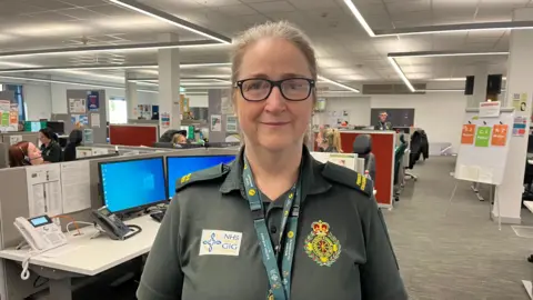 Getty Images Ambulance service worker Gill Pleming, wearing a dark green ambulance service polo shirt carrying the NHS Wales logo, smiles at the camera. Behind her, staff in the ambulance control room wear headsets and work at their screens