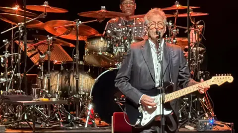 A man, Eric Clapton, stood holding a guitar while performing on a stage.