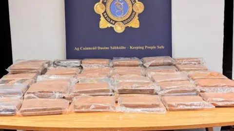 More than twenty vacuum packed brown packages lie on a table in front of a large navy sign, written on it the words 'Garda National Drugs and Organised Crime Bereau'. In the middle of the sign is the unit's insignia, a traditionally designed police badge with an ornate golden border. 