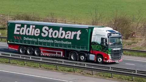 Getty Images A green, white and red truck driving on dual carriageway. It has 'Eddie Stobart Delivering Sustainable Distribution' in white writing. A field can be seen to the side of the road.