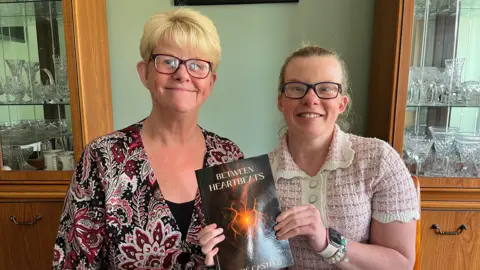 Zoë Castle and her mum Di sitting together in front of a cabinet filled with glass bowls and cups. Zoë has her hair tied back and is wearing glasses. She is holding up a book which is black with an orange heart in the centre. Di has short blonde hair and a floral patterned top. 
