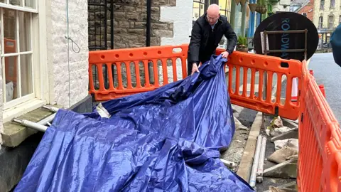Olivia Richwald/BBC A man is pulling a large blue tarpaulin across a hole in the road outside his pub.