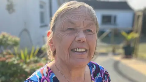 An elderly woman smiles and looks just off camera. She has mid length light hair. She is wearing a purple and blue patterned top. She is sitting in a garden.
