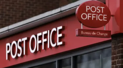 Getty Images Close-up of a Post Office storefront with red branding, featuring large white “POST OFFICE” lettering across the shop fascia. A circular red Post Office sign and a smaller “Bureau de Change” sign are mounted above the entrance, with brickwork and windows visible in the background.