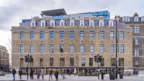 Royal Institute of British Architects The exterior of the Town Hall with people standing outside on the pavement at a set of traffic lights. There are four rows of vertical windows along the facade, the building is largely brown brick and the roof is slanted with grey tiles. 