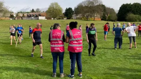Martin Heath/BBC A group of runners on a grass park. One man in a red and blue top is approaching the finish, where there are two volunteers wearing parkrun vests
