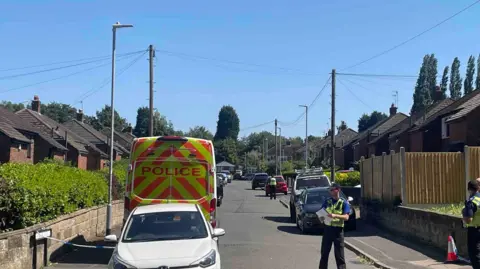 Police officers standing at the junction in a residential area. One officer is in the middle of the road holding some paper. Two others are standing on a pavement on the right. A parked police van is on the left and several other parked cars can be seen along the road.