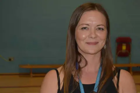 Suffolk New College Danielle is standing in the sports hall. The photo shows a head and shoulders image of her in a black sleeveless sports top. She has a blue lanyard around her neck.