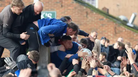 RKP Photography A group of men, some having climbed on a fence, wrestle over the ball