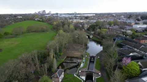 Aerial view of a canal and lock flight running through a green open space, with buildings and housing visible in the distance.