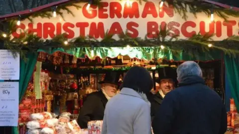 A biscuit stall at Lincoln Christmas Market showing two servers dressed in Victorian costume, including top hats. A sign, in large red letters, reads "German Christmas biscuits".