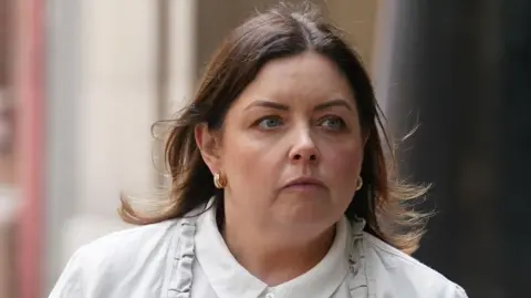 Deirdre Hargey, pictured from the shoulders up. She is wearing a white shirt and a white jacket and has long brown hair. 