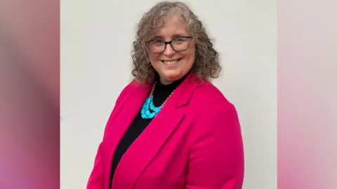 Supplied Anna Smith, who has shoulder-length brown and grey hair and is wearing glasses, smiles at the camera. She is wearing a pink jacket and top. 