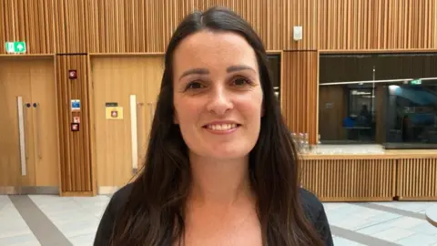 Kit Taylor/BBC Žena usmívající se do kamery, s dlouhými hnědými vlasy a černým topem. 