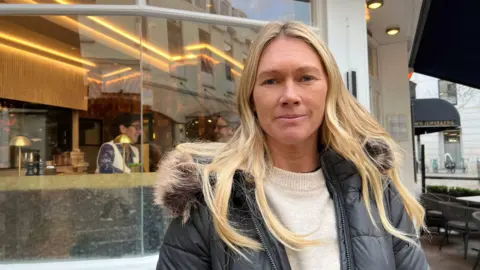 Debbie Vautier is wearing a black jacket and white jumper, and is standing outside. She has long blonde hair. 