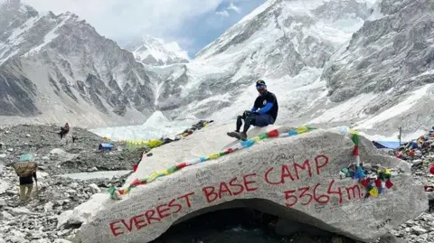 Hari Budha Magar Hari sits with his prostectic legs on a large rock which has the words Everest Base Camp sprayed onto it. Behind him are the mountains and some people who are starting their treks. There are colour ribbons on the rock as well.