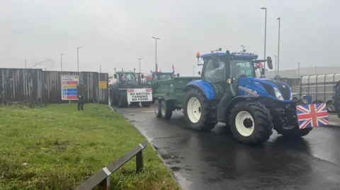 A blue tractor with a union flag on the front is parked in front of another tractor with a sign on the front reading "no British farming no food"