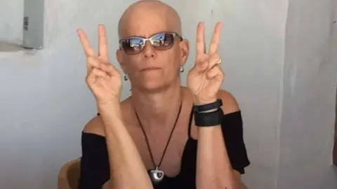 Alison Dalgliesh Nortje sat against a white wall. She has both hands up and is doing the 'peace' sign.