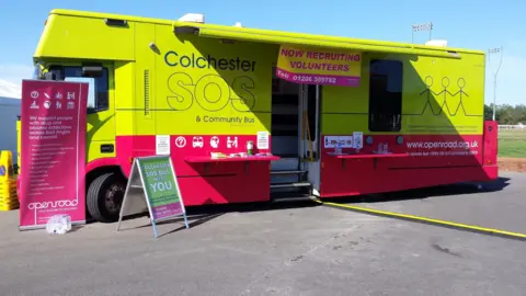 A lime green and pink bus with Colchester SOS written over it.
It is parked on tarmac floor.
There are posters and flyers outside of the bus.