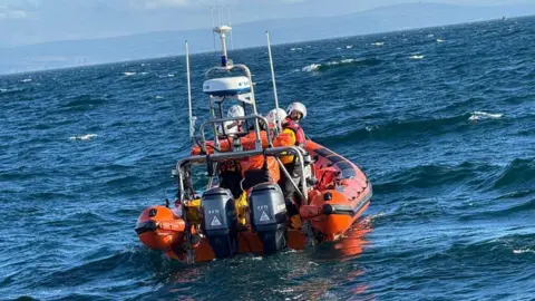 RNLI New Quay Orange life boat with crew wearing helmets on board seen in the sea from behind with engines at the back