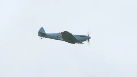 A Spitfire painted in a pale blue colour with a circular logo on the side flies through the air.