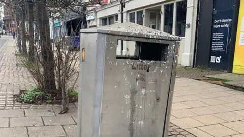 A silver metal litter bin which appears to be dirty and is covered in stickers.