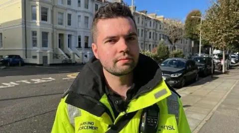 Finn Gregory is wearing his high vis uniform and is standing on the pavement of a street in Hove, with a number of parked cars behind him.