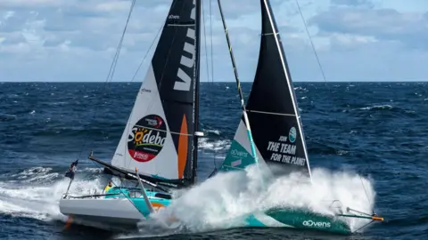 Pierre Bouras A sailaing boat going fast with spray with the ocean around it and a man in a  yellow sailing outfit in the cockpit.