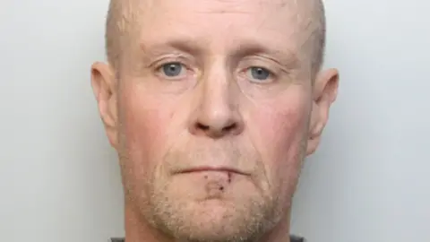 Darren Morris looks into the camera for his custody image. He has a small cut to his lip. He has short facial hair, which is light grey and ginger-y, and short hair of the same colour.