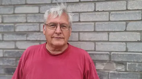 PA Media A head and shoulder shot of Prof David Martill, taken against a grey brick wall. He has short, white hair, and wears thin-rimmed rectangular glasses and a red T-shirt
