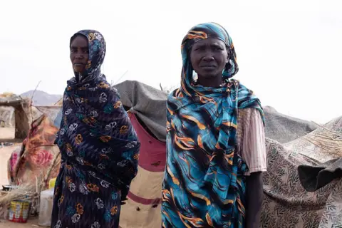 AFP via Getty Images Two women stand in front of their makeshift shelter in the Um Yanqur camp, located on the southwestern edge of Tawila.