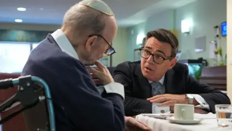 PA Media On the left is a man sat in a wheelchair. He is wearing a blue jumper, a white shirt, glasses and a kippah. To his right is Andy Burnham, who is also sat down with his arms placed on a table. Burnham is wearing a dark-coloured suit jacket, white shirt and a tie. The two men are in discussion. This is taking place in a care home.